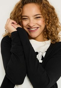Cardigan noir à manches longues, porté sur un sweat-shirt de couleur claire avec un motif graphique. Texture douce, coupe ajustée, look décontracté.