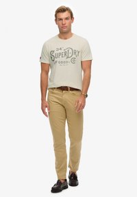 Camiseta de algodón beige con gráfico "Superdry", pantalones chinos color tan y zapatos marrón oscuro. El modelo está de pie en una pose neutral sobre un fondo liso.