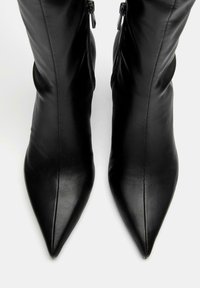 Fekete, vádlit takaró csizmák sima bőrből, hegyes orral és oldalsó cipzárokkal az elengedéshez, melyek karcsú, testhezálló sziluettet mutatnak.