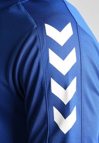 Gros plan sur la manche d'une chemise bleue avec quatre flèches blanches en chevron pointant vers le bas le long du bras.