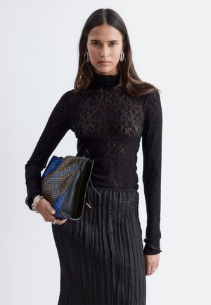 Jeune femme aux longs cheveux foncés portant un haut en dentelle noire et une jupe plissée, tenant un grand sac à main bleu et marron à motifs.