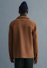 Brauner Wollmantel mit Kragen, Knöpfen an den Manschetten und einer Aufhängeschlaufe. Kombiniert mit dunkelgrünen Hosen und einer marineblauen Mütze.