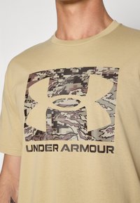 Camiseta de manga corta en color beige con un gran logo de camuflaje y el texto "UNDER ARMOUR" en negrita negra en la parte inferior.