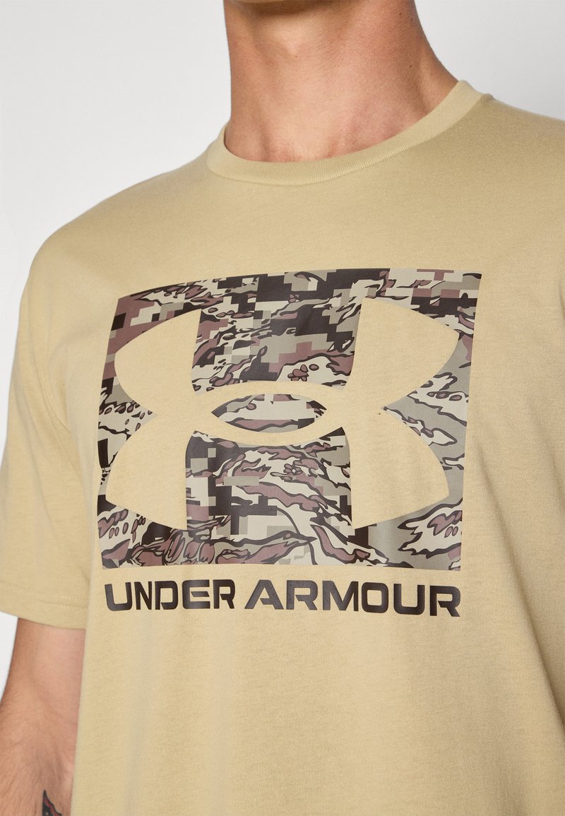 Camiseta de manga corta en color beige con un gran logo de camuflaje y el texto "UNDER ARMOUR" en negrita negra en la parte inferior.