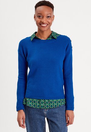 Pull bleu avec un motif texturé, col rond, manches longues. En dessous, une chemise verte à motifs est visible. Porté avec un jean en denim.