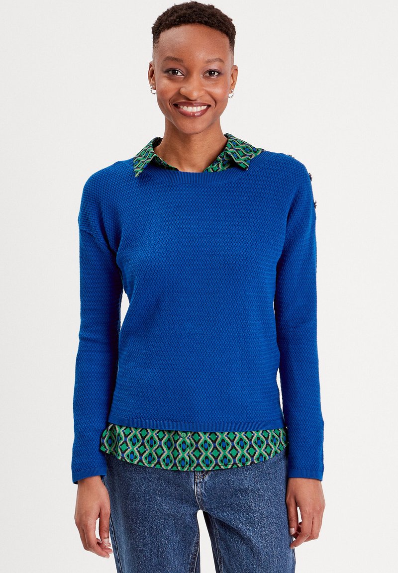 Pull bleu avec un motif texturé, col rond, manches longues. En dessous, une chemise verte à motifs est visible. Porté avec un jean en denim.