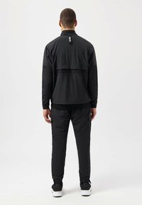 Black athletic jacket with mesh panels, high collar, and logo tag at back, paired with matching black pants and sporty shoes.