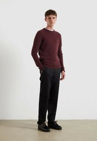 Maglione in tessuto a maglia color bordeaux con collo rotondo, maniche a coste e un piccolo logo, abbinato a pantaloni neri a sigaretta e calzature scure.