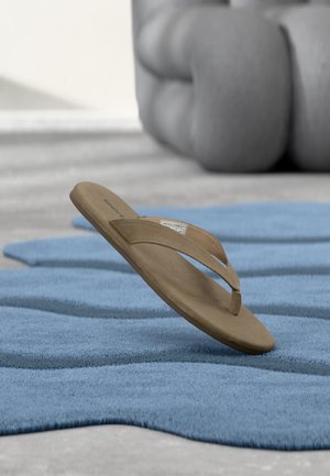 Bruine teenslipper hangend boven een blauw gestructureerd tapijt op een lichte vloer, met een vervaagd groot grijs object op de achtergrond.