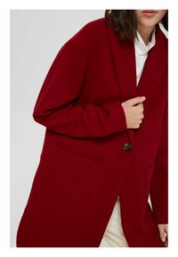 Czerwony wełniany płaszcz z długimi rękawami, dużą przednią kieszenią i zapięciem na guziki. Posiada kołnierz szalowy i luźny krój.