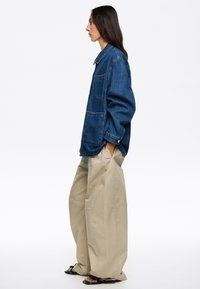 Giacca di jeans oversize in blu scuro, con taschino sul petto e chiusura a bottoni. Abbinata a pantaloni larghi color crema.