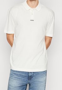Camisa polo blanca de tejido suave, con cuello, mangas cortas y un logo negro de "HUGO" centrado en el pecho. Usada con vaqueros azules.