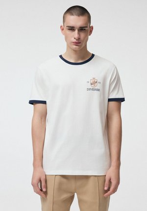 Młody mężczyzna ubrany w białą koszulkę z granatowym kołnierzykiem i krawędziami rękawów, beżowe spodnie oraz mały herb z napisem "DIVERSE 1993" na piersi.