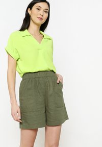 Neon žalios spalvos trumpųjų rankovių blusas su V formos iškirpte, derinama su alyvuogių žalios spalvos, laisvo kirpimo šortais, turinčiais elastingą liemenį ir šonines kišenes.