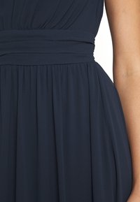 Námořnické modré šaty, vyrobené z měkké, splývavé tkaniny s plisovaným designem. Mají pásek, který vytváří na sukni nařasenou texturu.