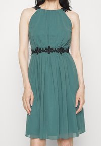 Turkos halterneck klänning med åtsittande midja, plisserad kjol och svart blommönster över bysten och midjan. Lätt och svävande tyg.