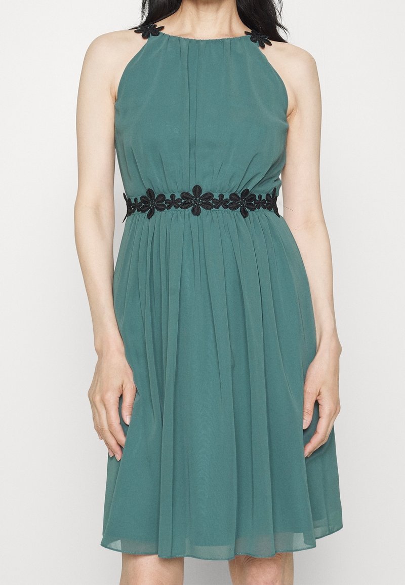 Turkos halterneck klänning med åtsittande midja, plisserad kjol och svart blommönster över bysten och midjan. Lätt och svävande tyg.