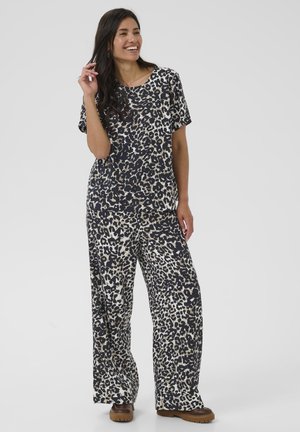 Mujer sonriente posando con conjunto a juego de top de manga corta y pantalones de pierna ancha con estampado de leopardo negro y beige y zapatos marrones sobre fondo gris.