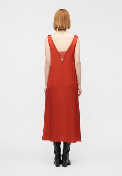 Κόκκινο αμάνικο φόρεμα με βαθύ V στην πλάτη και δαντελένια λεπτομέρεια, κατασκευασμένο από απαλό ύφασμα, που φτάνει μέχρι τη μέση της γάμπας, συνδυασμένο με μαύρες μπότες μέχρι το γόνατο.