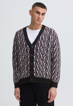 Uomo con capelli corti indossa un cardigan con bottoni bordeaux, nero e bianco sopra una t-shirt bianca e pantaloni neri su uno sfondo semplice.