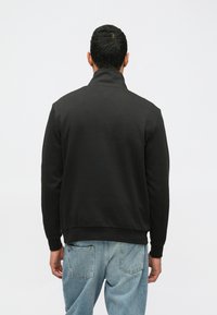 Felpa nera con colletto alto, tessuto testurizzato, polsini e orlo a coste, indossata sopra jeans blu scoloriti, vista da dietro.