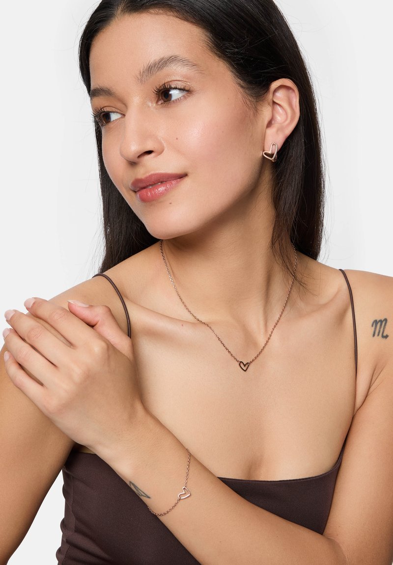 Set di gioielli in oro rosa con una collana a pendente a forma di cuore, orecchini coordinati e bracciale, indossati da un modello con pelle liscia.