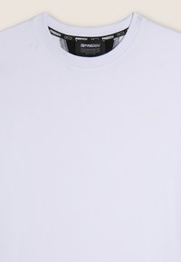 Camiseta de algodón blanca con escote redondo, que presenta un sutil logo en relieve en amarillo claro en el frontal y una etiqueta negra en el interior del cuello.