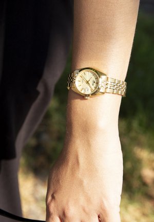 Goldfarbene Armbanduhr mit rundem Ziffernblatt, metallischem Armband und beigem Zifferblatt, das Stundenmarker und ein Datumsfenster zeigt, auf einem Handgelenk platziert.