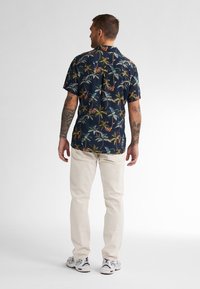 Camicia a maniche corte blu navy con motivo a palma, abbinata a pantaloni beige chiaro e sneakers. La camicia presenta un colletto e una vestibilità rilassata.