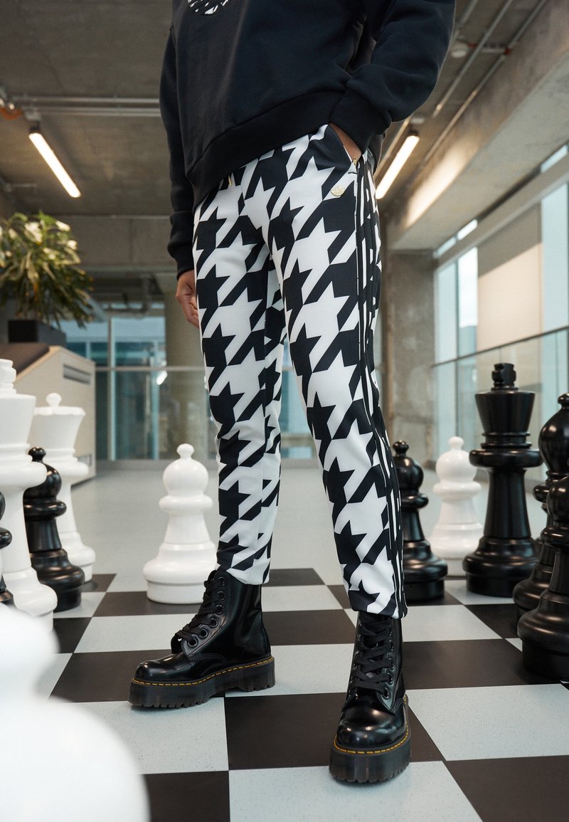 Černobílé joggery s kostkovaným vzorem houndstooth a bočními pruhy, doplněné lesklými černými šněrovacími botami a černou mikinou. V pozadí šachové figurky.