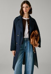Marineblauwe trenchcoat met een dubbelbreasted ontwerp, gecombineerd met een bruine cardigan, witte blouse en brede blauwe jeans. Houdt een bruine leren tas vast.