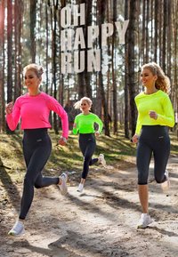 Tre donne corrono su un sentiero di terra in una foresta. Indossano magliette a maniche lunghe di un luminoso colore rosa e verde, con leggings neri a vita alta e scarpe da ginnastica.