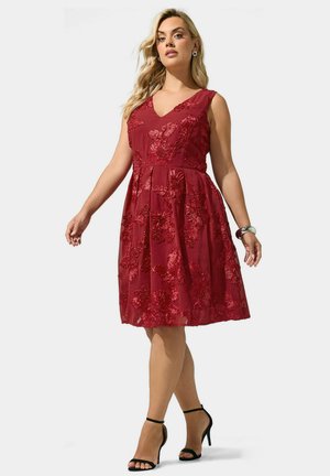 Mujer vestida con un vestido rojo sin mangas bordado con flores, de escote en V y sandalias negras de tacón alto, posando con los brazos ligeramente extendidos.