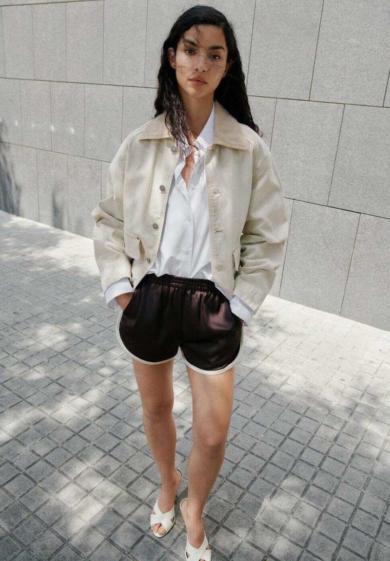 Beige cropped jack met een witte blouse, zwarte satijnen shorts met witte biezen, en witte sandalen, tegen een gestructureerde grijze achtergrond.