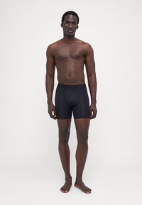 Fekete boxer alsónadrág, rugalmas anyagból készült, egyszínű, elasztikus derékrésszel. A modell mezítláb áll egy sima háttér előtt.
