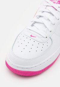 Valkoinen nahkakenkä vaaleanpunaisella kumipohjalla, rei'itetyllä varpaalla ja valkoisilla nauhoilla. Sivussa on pieni vaaleanpunainen Nike-logo.