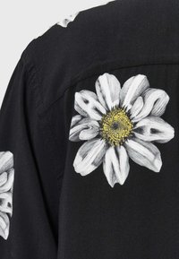 Czarna koszula z dużymi białymi nadrukami stokrotek o żółtych środkach. Tekstura wydaje się gładka, z casualowym krojem i detalami w postaci guzików.