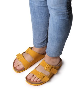 Sandales en suède jaune avec deux boucles ajustables, semelle en liège et semelle extérieure en caoutchouc texturé, portées avec un jean bleu.