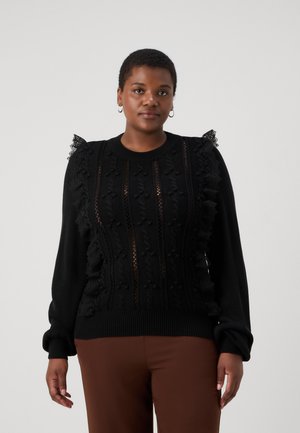 Svart stickad tröja med texturerade mönster, spetsdetaljer på axlarna och ribbade ärmslut. Matchas med bruna byxor.