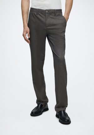 Homme portant un pantalon de costume gris foncé avec un bouton et une fermeture éclair, une main dans la poche, associé à des chaussures noires sans lacets.
