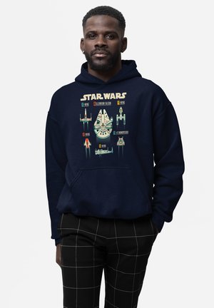 Mężczyzna noszący granatową bluzę z kapturem z ilustracją statków Star Wars i etykietami, w połączeniu z czarnymi, kratowanymi spodniami, stojący na białym tle.