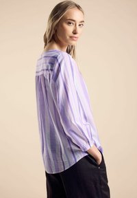 Camicia leggera a maniche lunghe di colore lavanda con strisce verticali di diverse tonalità. Presenta un retro arriccito e una vestibilità rilassata. Abbinata a pantaloni neri.