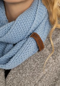 Lichtblauwe gebreide sjaal met een gestructureerd patroon. Voorzien van een bruine leren label met "Knit Factory." Draag over een grijze harige jas.
