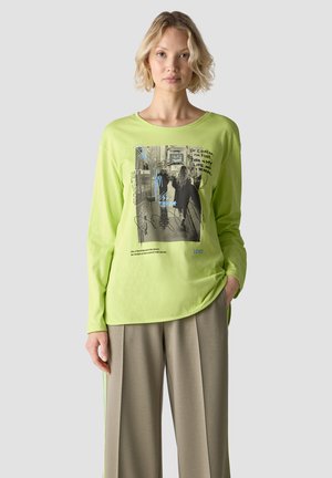 Frau mit kurzen blonden Haaren trägt ein limonengrünes Langarmshirt mit Grafikdruck und beige weite Hosen, steht vor einem einfarbigen Hintergrund.