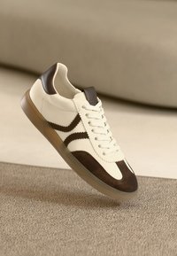 Valged tossud tumepruuni seemisnahast detailidega, tekstuurse pinnaga, lamedate paeltega ja läbipaistvast kummist tallaga. Küljed kaunistatud chevron-mustriga.