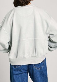 Sudadera gris claro de gran tamaño con cuello redondo, ribete acanalado en el bajo y hombros caídos, combinada con jeans azules, que muestran un ajuste relajado.
