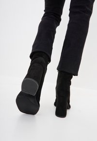 Person wearing black suede heeled ankle boots and dark pants, walking on a smooth white surface.