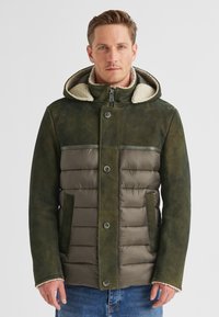 Mann in einer grünen gepolsterten und Wildleder-Winterjacke mit Kapuze und Lammfell-Futter, der vor einem schlichten hellen Hintergrund steht.