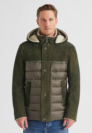 Mann in einer grünen gepolsterten und Wildleder-Winterjacke mit Kapuze und Lammfell-Futter, der vor einem schlichten hellen Hintergrund steht.