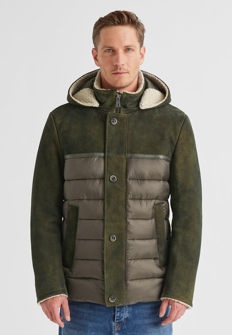 Mann in einer grünen gepolsterten und Wildleder-Winterjacke mit Kapuze und Lammfell-Futter, der vor einem schlichten hellen Hintergrund steht.
